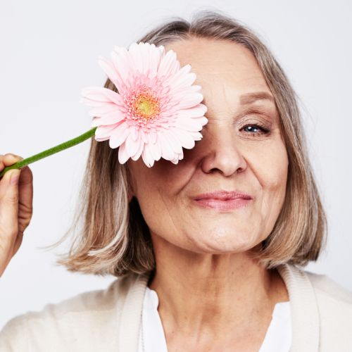 Mulher mais velha sorrindo segurando uma flor rosa vibrante na frente de um olho